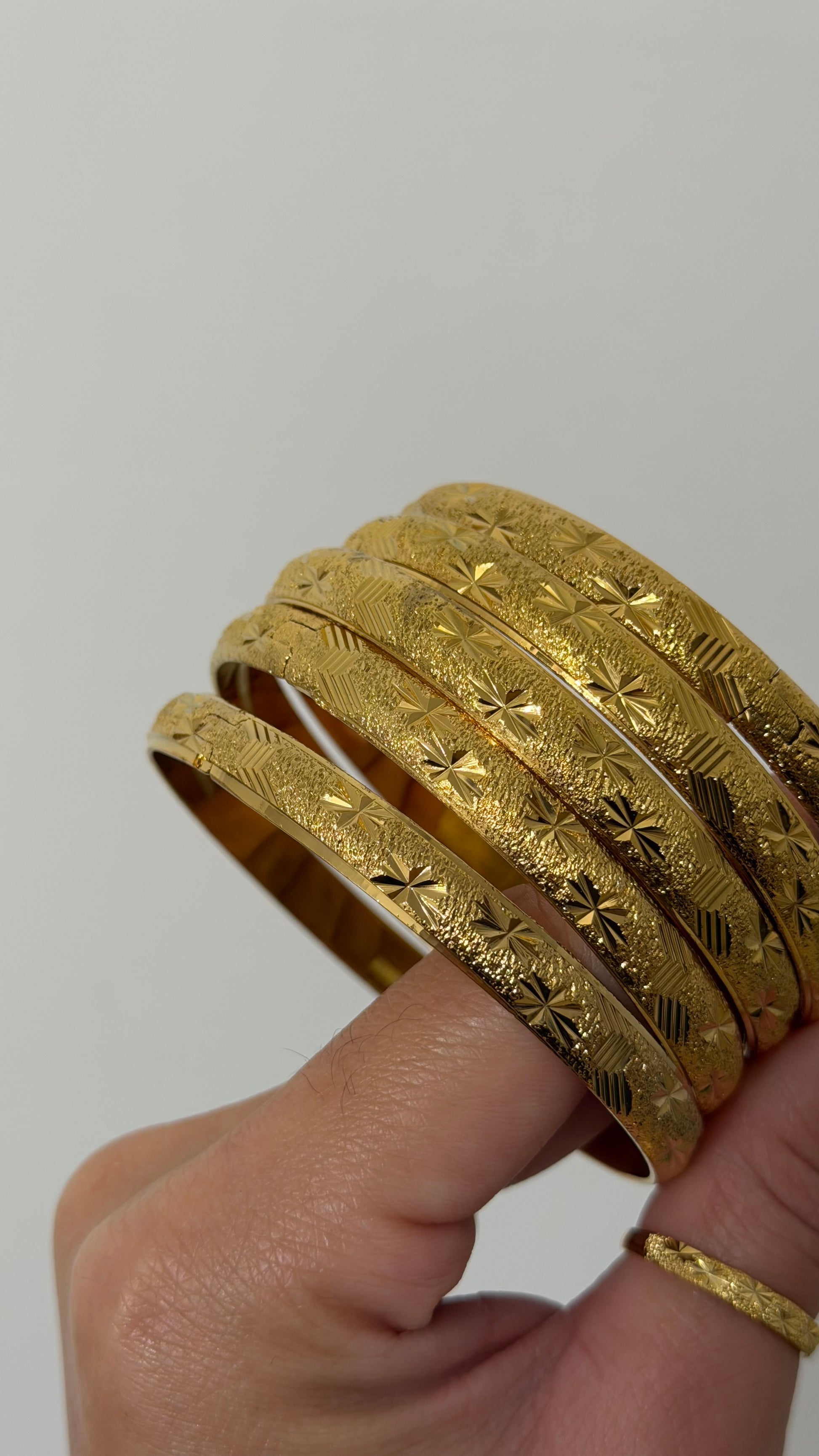 A hand holding a stack of five textured gold bangles with engraved floral and geometric patterns, set against a plain light gray background.