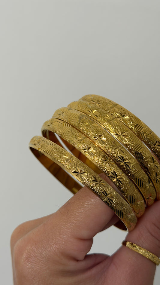A hand holding a stack of five textured gold bangles with engraved floral and geometric patterns, set against a plain light gray background.